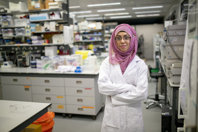 A portrait young female biochemist in a lab, standing tall and looking proud in her white lab coat and traditional hijab.